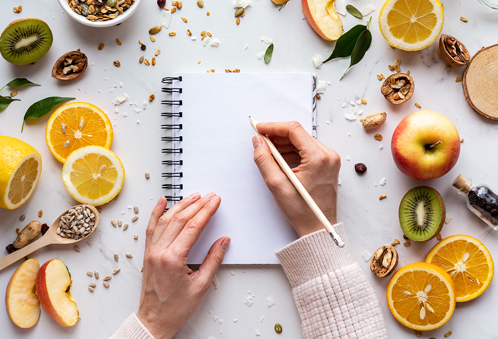Female hand write in notebook on healthy food background, women diet nutrition recipe menu, fresh summer fruit granola seeds on white table organic super food, health care detox, meal planning