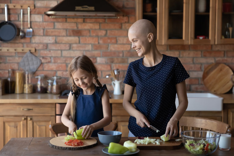 Oncology care. Smiling young sick cancer patient bald hairless mom and little daughter cooking together at home kitchen, happy ill mother and small girl child make healthy diet food together, vegetarian concept
